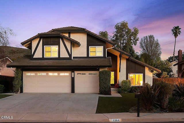 tudor-style house with concrete driveway, an attached garage, fence, and stucco siding