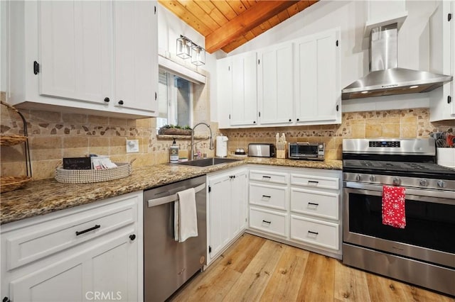 kitchen with wall chimney exhaust hood, appliances with stainless steel finishes, vaulted ceiling with beams, white cabinetry, and a sink