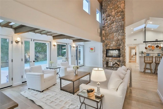 living room featuring a skylight, light wood-style flooring, beamed ceiling, a stone fireplace, and french doors