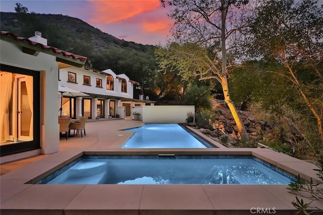 pool at dusk featuring a mountain view, an outdoor pool, a patio, and an in ground hot tub