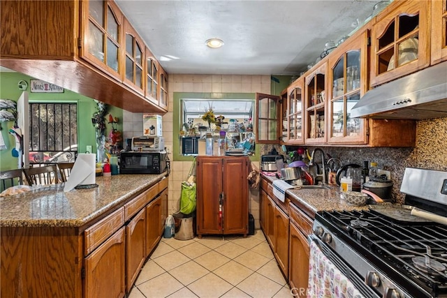 kitchen featuring stainless steel range with gas stovetop, brown cabinets, ventilation hood, and glass insert cabinets