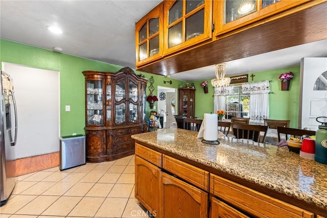 kitchen with light tile patterned floors, stainless steel fridge, glass insert cabinets, and brown cabinetry