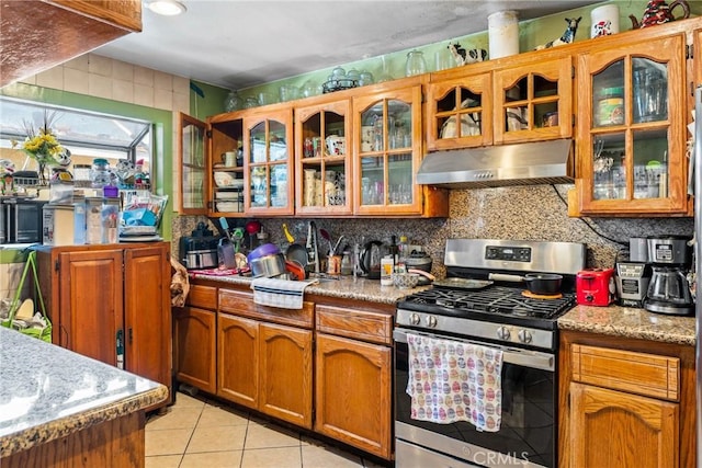 kitchen with under cabinet range hood, backsplash, gas stove, light tile patterned flooring, and brown cabinetry