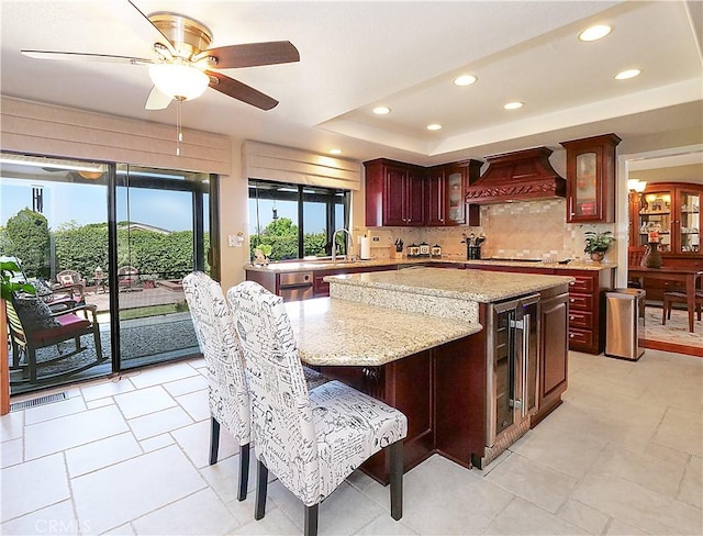 kitchen with a tray ceiling, custom exhaust hood, tasteful backsplash, a sink, and beverage cooler