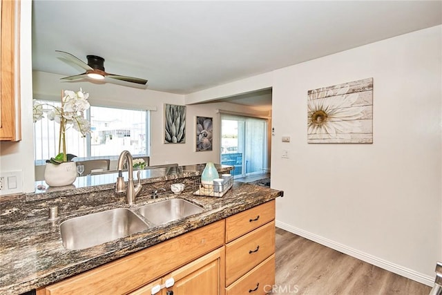 kitchen featuring light wood finished floors, a ceiling fan, a sink, dark stone countertops, and baseboards