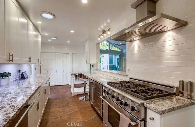 kitchen with decorative backsplash, light stone counters, high end stainless steel range, wall chimney range hood, and white cabinetry