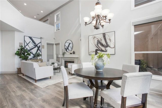 dining room featuring recessed lighting, a warm lit fireplace, light wood-style floors, a towering ceiling, and a notable chandelier