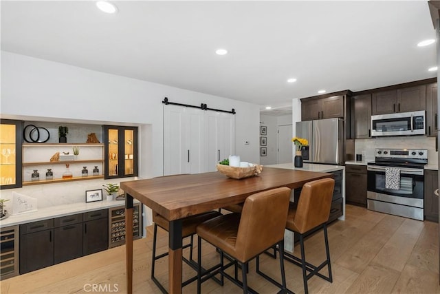 kitchen with a barn door, dark brown cabinetry, appliances with stainless steel finishes, light wood-type flooring, and backsplash