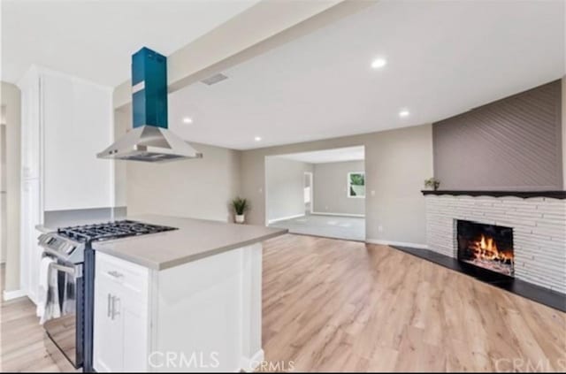 kitchen featuring white cabinets, a stone fireplace, light wood-style floors, stainless steel range with gas stovetop, and exhaust hood