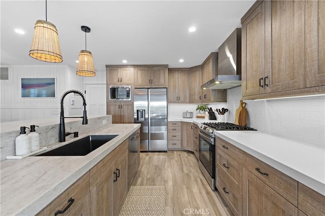 kitchen with light wood-style flooring, stainless steel appliances, a sink, wall chimney range hood, and decorative light fixtures