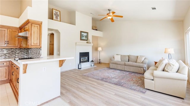 living room featuring light wood finished floors, arched walkways, a towering ceiling, a fireplace with flush hearth, and ceiling fan