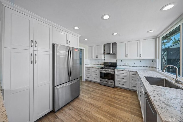 kitchen featuring light wood finished floors, stainless steel appliances, white cabinetry, a sink, and wall chimney exhaust hood