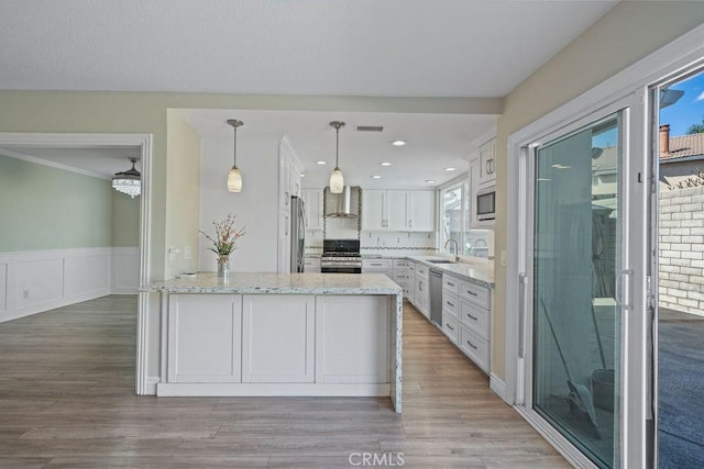 kitchen with white cabinets, appliances with stainless steel finishes, light stone counters, a peninsula, and wall chimney range hood