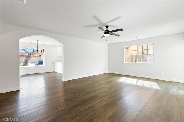 unfurnished living room with dark wood-type flooring, arched walkways, baseboards, and ceiling fan with notable chandelier