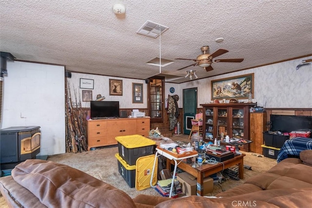 carpeted living area featuring a textured ceiling, visible vents, a ceiling fan, wainscoting, and a wood stove