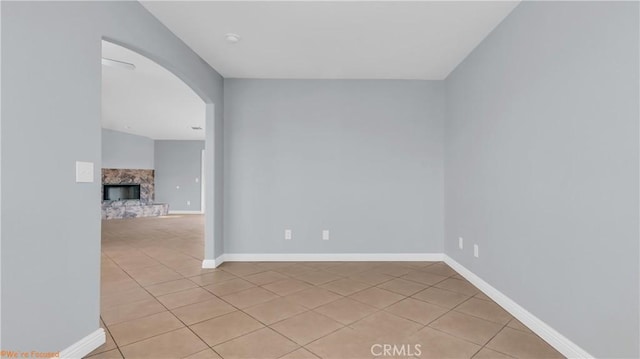 spare room featuring arched walkways, a fireplace with raised hearth, light tile patterned floors, and baseboards