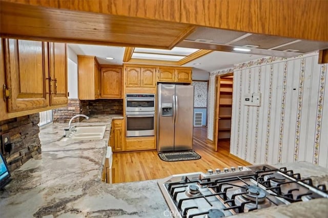 kitchen featuring light wood-style flooring, appliances with stainless steel finishes, brown cabinetry, a sink, and light stone countertops