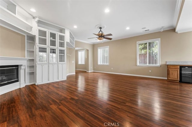 unfurnished living room with crown molding, wine cooler, dark wood finished floors, a glass covered fireplace, and a ceiling fan