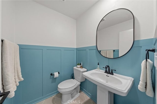 bathroom featuring a wainscoted wall, a decorative wall, toilet, a sink, and tile patterned flooring