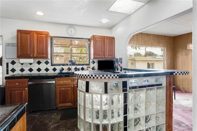 kitchen with tile countertops, wooden walls, brown cabinetry, dishwasher, and backsplash