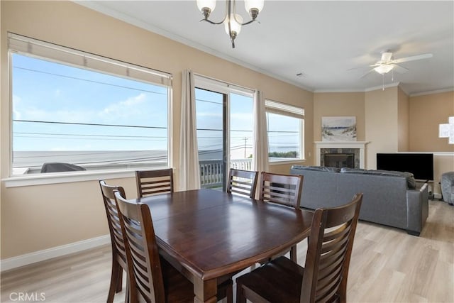 dining space featuring baseboards, light wood finished floors, ornamental molding, a glass covered fireplace, and ceiling fan with notable chandelier