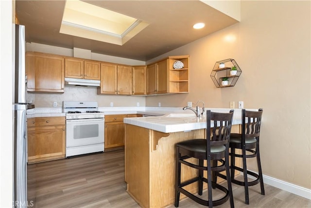 kitchen featuring tile counters, under cabinet range hood, a breakfast bar area, white range with gas stovetop, and a peninsula