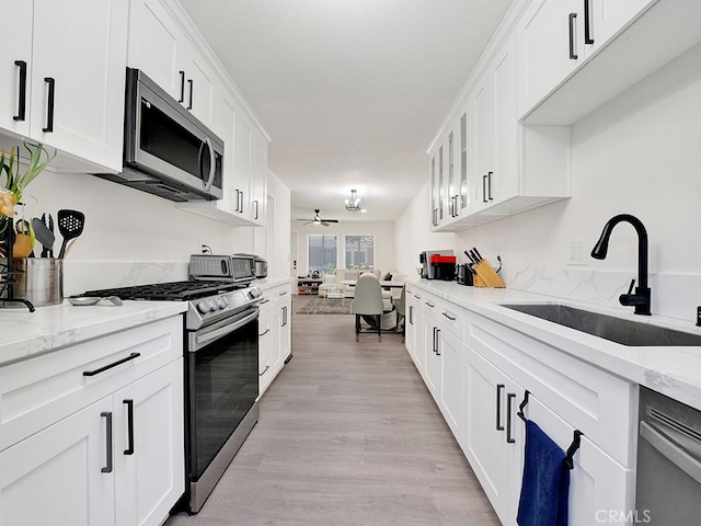 kitchen with light wood finished floors, stainless steel appliances, white cabinets, a sink, and ceiling fan