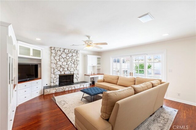 living area with baseboards, a ceiling fan, dark wood-style floors, a stone fireplace, and built in shelves