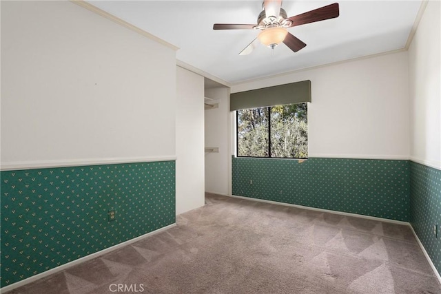 carpeted empty room featuring wallpapered walls, a ceiling fan, crown molding, and wainscoting