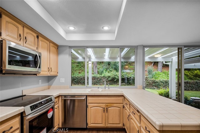 kitchen with tile countertops, a peninsula, a sink, stainless steel appliances, and a raised ceiling