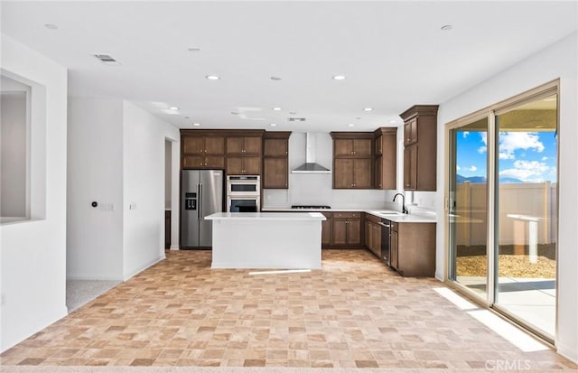 kitchen with light countertops, appliances with stainless steel finishes, a sink, wall chimney range hood, and a kitchen island