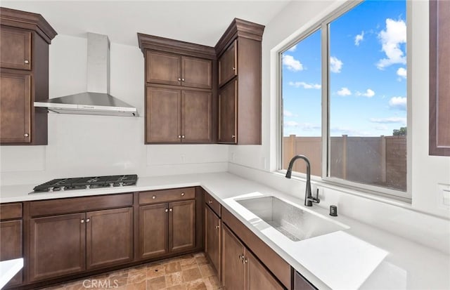 kitchen with light countertops, stainless steel gas stovetop, a sink, wall chimney range hood, and dark brown cabinetry