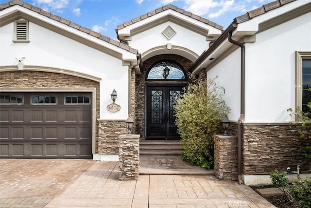 entrance to property with decorative driveway, stone siding, an attached garage, and stucco siding