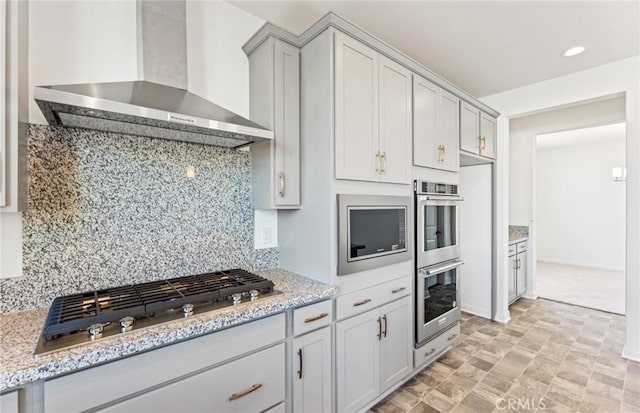 kitchen with light stone countertops, wall chimney exhaust hood, appliances with stainless steel finishes, and tasteful backsplash