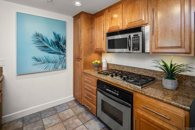 kitchen with appliances with stainless steel finishes, dark stone counters, brown cabinetry, and baseboards