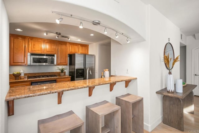 kitchen featuring brown cabinetry, appliances with stainless steel finishes, a breakfast bar, a peninsula, and light stone countertops