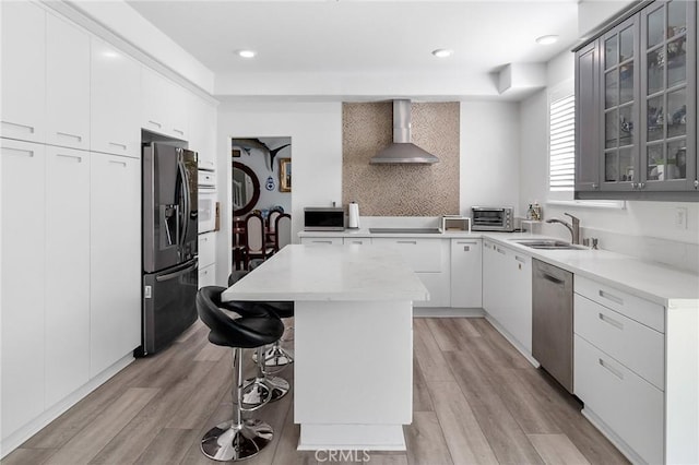 kitchen featuring a sink, a kitchen island, appliances with stainless steel finishes, wall chimney range hood, and glass insert cabinets