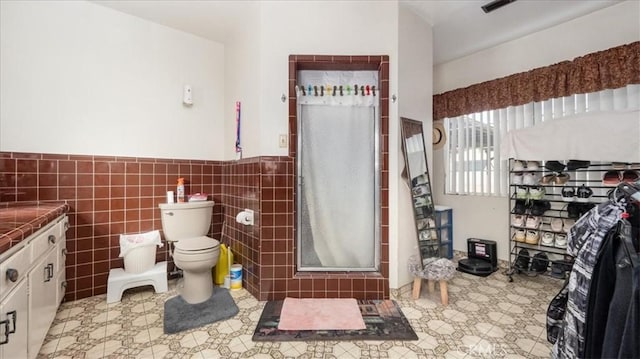 bathroom with a stall shower, toilet, a wainscoted wall, vanity, and tile walls