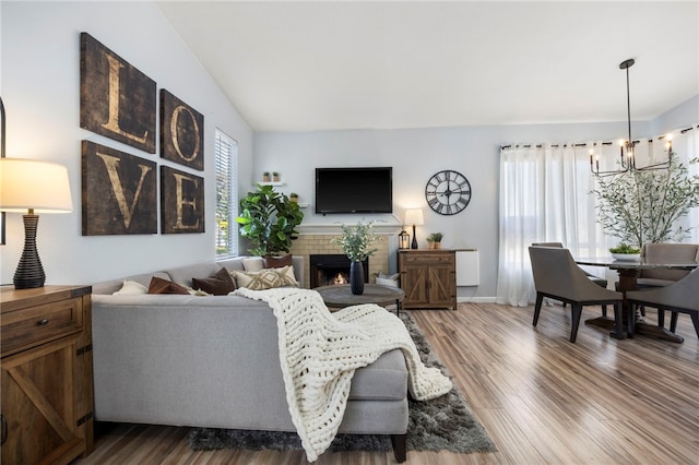 living area featuring vaulted ceiling, a lit fireplace, wood finished floors, and an inviting chandelier