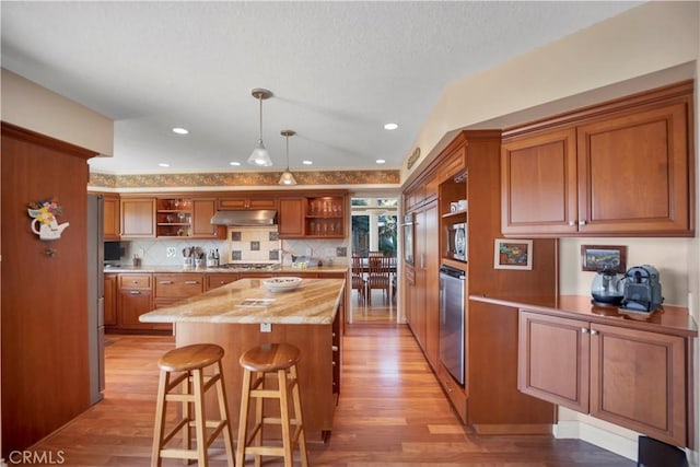 kitchen featuring brown cabinetry, a center island, stainless steel appliances, under cabinet range hood, and open shelves