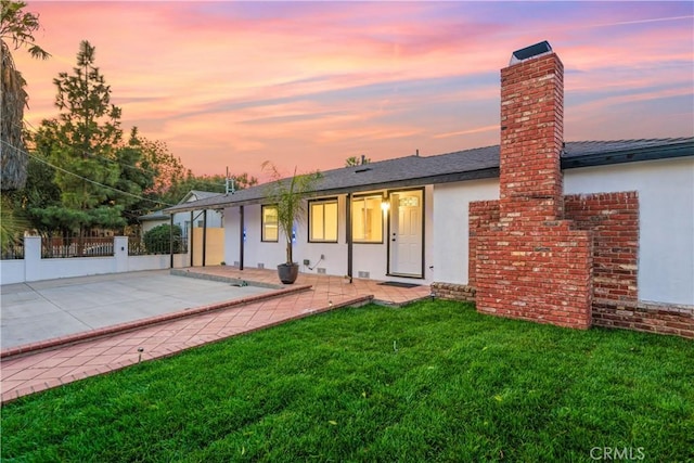 back of house at dusk with a patio, a shingled roof, fence, a lawn, and stucco siding