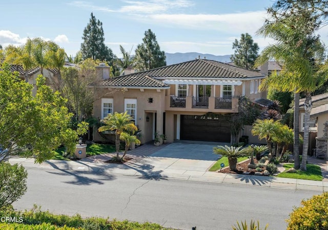 mediterranean / spanish house featuring an attached garage, a balcony, driveway, a tiled roof, and stucco siding