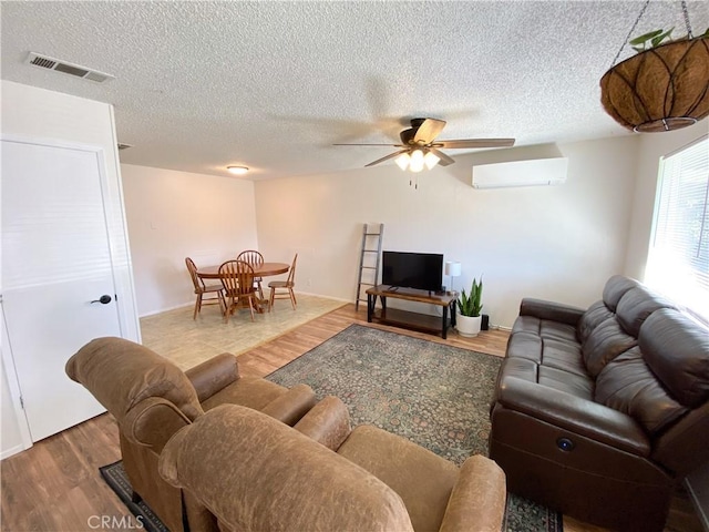 living room featuring a wall unit AC, wood finished floors, visible vents, ceiling fan, and a textured ceiling