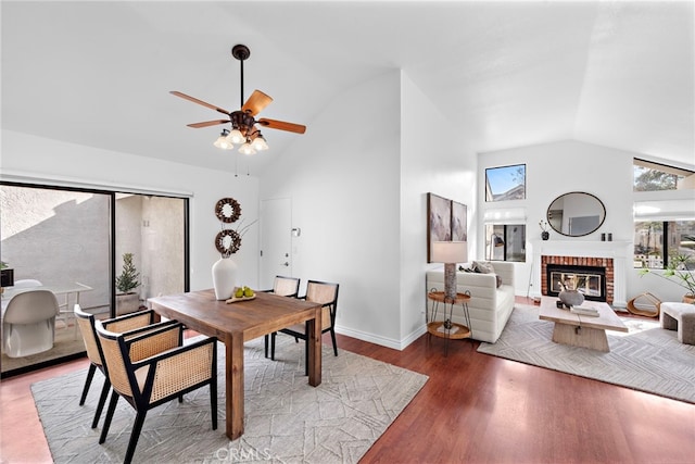 dining room with ceiling fan, high vaulted ceiling, a fireplace, wood finished floors, and baseboards