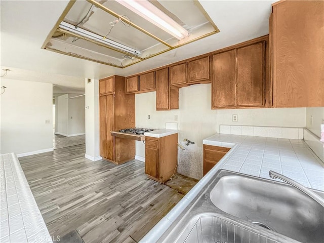 kitchen featuring baseboards, tile counters, brown cabinetry, wood finished floors, and a sink