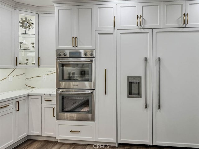 kitchen with double oven, light stone counters, dark wood-style floors, decorative backsplash, and glass insert cabinets