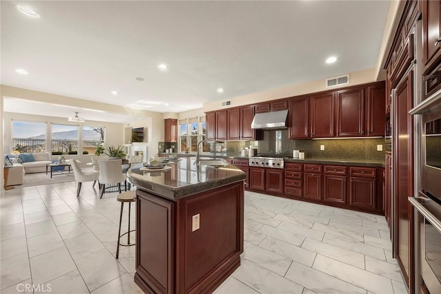 kitchen with reddish brown cabinets, stainless steel appliances, visible vents, under cabinet range hood, and a kitchen bar