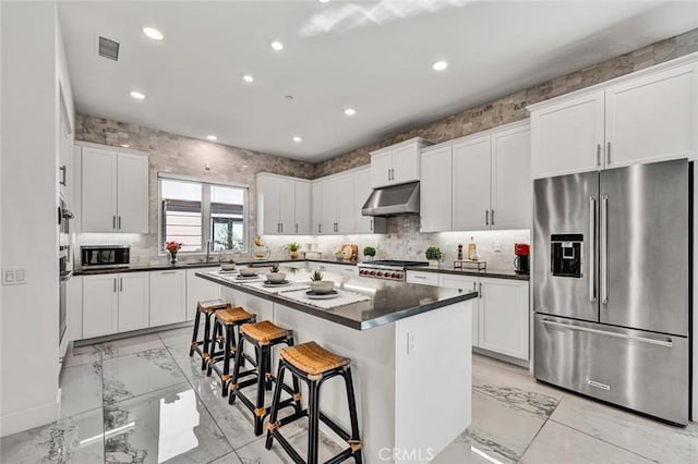 kitchen with under cabinet range hood, stainless steel appliances, marble finish floor, a center island, and dark countertops