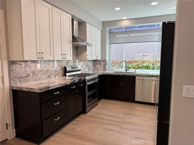 kitchen featuring a sink, backsplash, white cabinetry, stainless steel appliances, and wall chimney exhaust hood