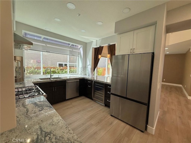 kitchen with wall chimney range hood, wine cooler, light stone counters, stainless steel appliances, and white cabinetry